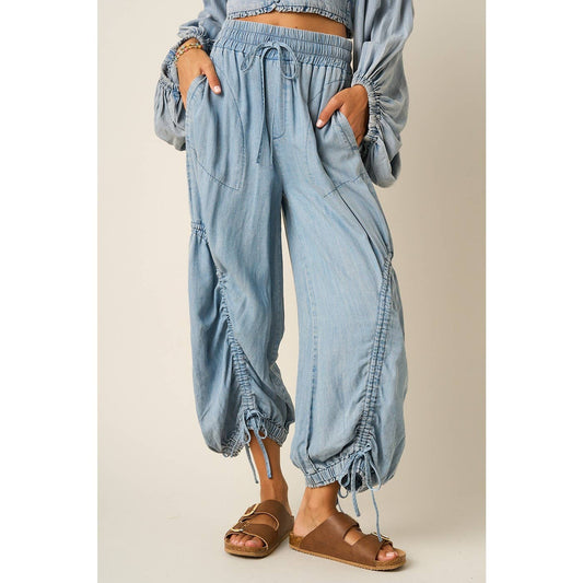 Light blue denim harem-style pants with drawstring waist and gathered cuffed hems, shown on a model wearing brown slide sandals against a plain beige studio background.