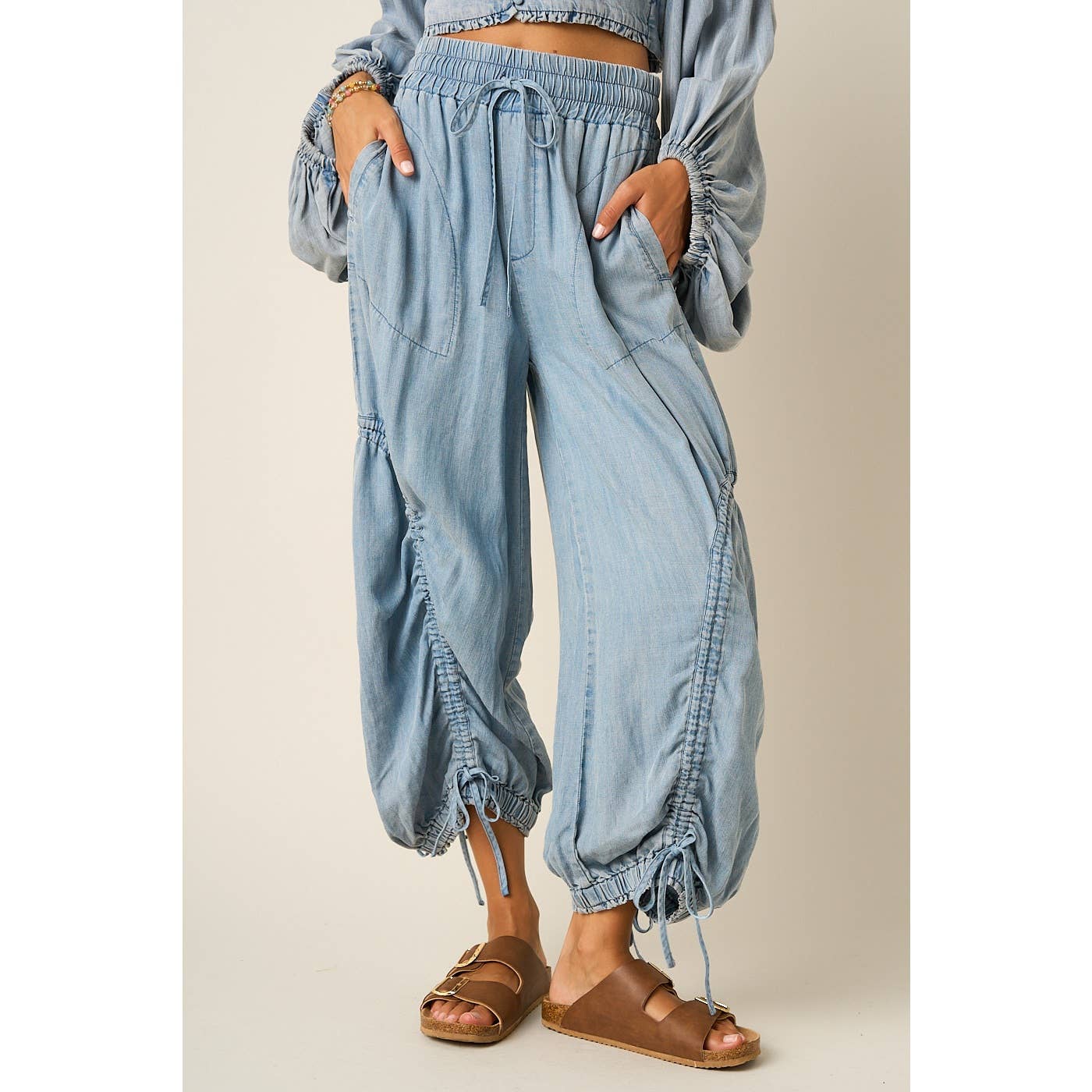 Light blue denim harem-style pants with drawstring waist and gathered cuffed hems, shown on a model wearing brown slide sandals against a plain beige studio background.